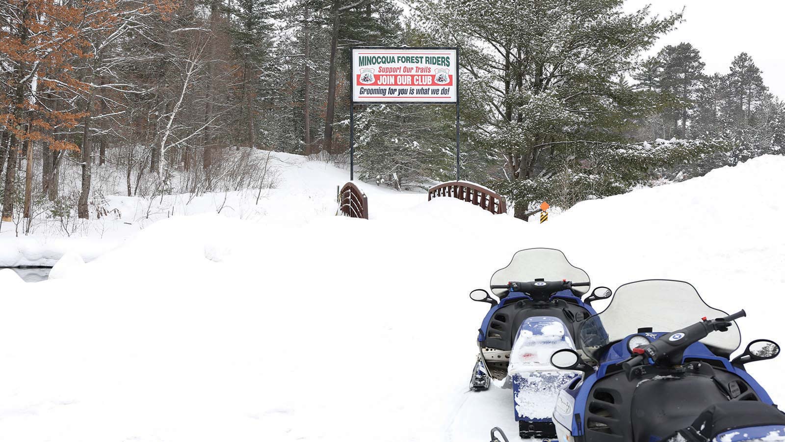 Two snowmobiles on front of a Minocqua Forest Riders trail sign on the snow covered trail
